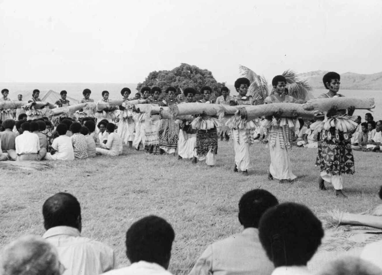 Multimedia Performance. Thirteen women in a line ceremoniously carry rolled up textiles through a seated crowd. The women wear richly decorated costumes and elaborate face paint, and those that are visible in this image all wear their hair in similarly short styles. Each carries a large, rolled-up textile horizontally and they walk in a neat line, gazing straight ahead. They are walking past small groups of people, seated cross-legged on the ground. 
