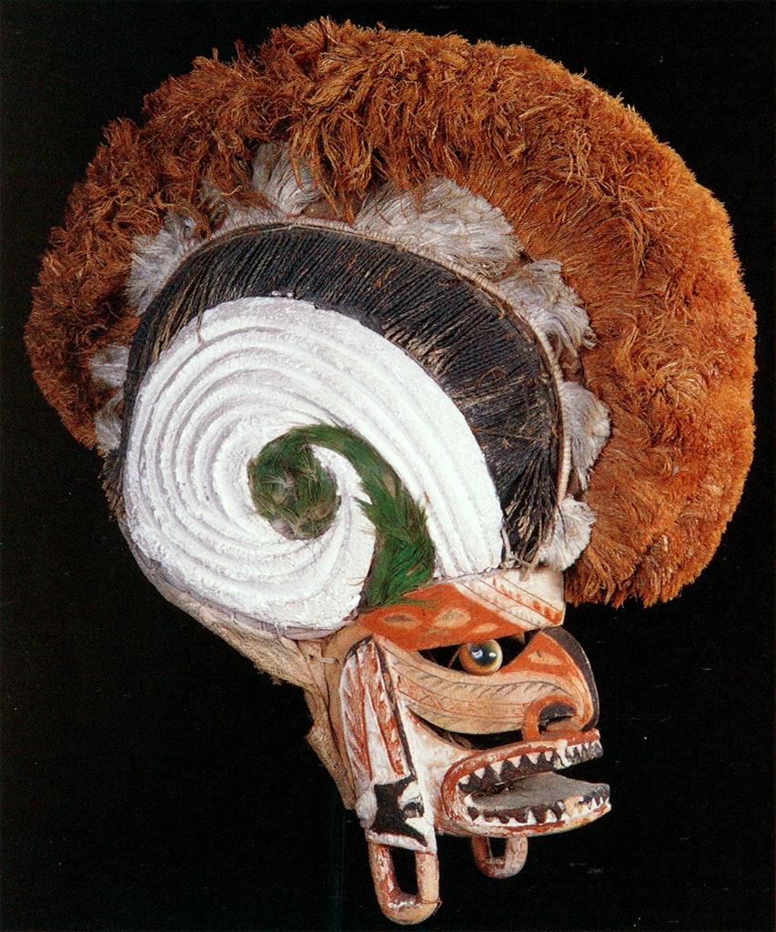 Mask. Painted wood, shells, and fiber form a fierce, human-like face, a large crest extending over the top of the head.
The painted wood of this mask describes an abstracted, human face with a gaping mouth full of sharply-pointed teeth. The dominant colors on the face are oranges, browns, and white, with some black accents in the eyes and on the long ear lobes.swirling patterns create the impression of the profile of a nose with a prominent black nostril. There is space cut out for the eyes, with a round, painted object for an eyeball fixed in the center of the opening. On the top of the head is a crest, almost rooster-like, forming a crescent-shaped headdress that is several times larger than the lower portion of the head. Green fibers and painted wood form the center of the crest, with orange fibers extending along the top of the crest.

