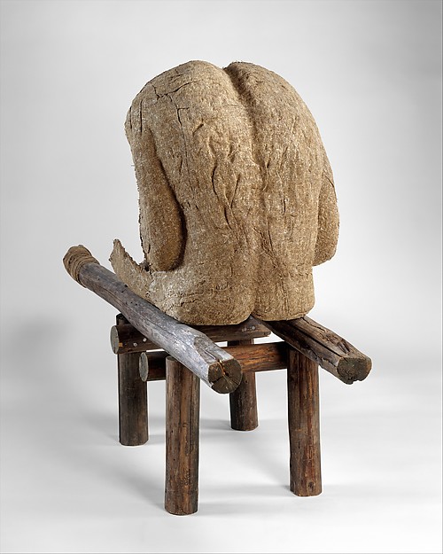 Sculpture. A roughly-textured, headless, human-sized figure sits, hunched over, on a wooden stretcher.
The materials used in this sculpture are burlap, wood, resin, nails, and string, resulting in a monochromatic brown color all over, though the wooden bench is a slightly deeper brown than the burlap figure. This view from behind the headless figure reveals a long, broad back with a deep crease between the shoulder blades. It appears to be missing most of its legs. The arms are bent as if the figure is leaning on its elbows, tucked tightly against the sides of the torso.

