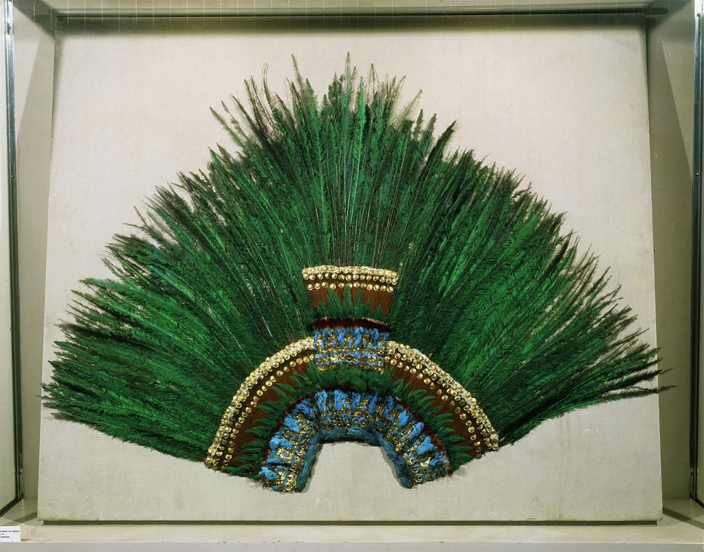 Headdress. A fan-shaped headdress is made of gold, with long, bright green and blue feathers spreading outwards.
The long, brilliant, blue and green feathers fan outwards and appear to shimmer in the light. In the center of the headdress, the base of the feathers are attached to three semi-circular rings of gold. Inside this series of rings is a deep red ring, inside of that is a pattern of gold, and small, bright blue feathers.

