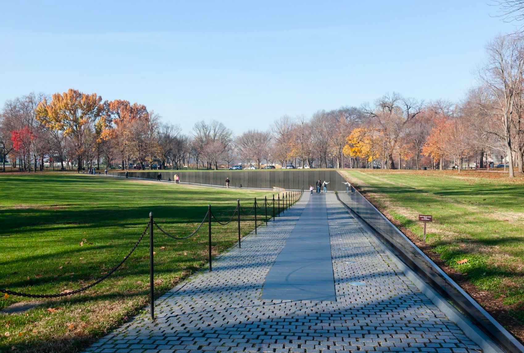 Installation. A permanent pathway stretches into the distance, bending sharply to the left, grass on both sides.
On the horizon are trees with fall-colored leaves, placing this constructed path in a park-like setting. Down the left side of the path is a simple barrier of posts and chains. Down the right side is a smooth, reflective wall that increases in height as the path slants downward, more deeply into the earth. There is writing on the wall in regular rows.

