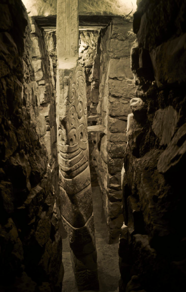 Architectural Sculpture. Carving of a larger-than-life, half-human, half-animal figure within a narrow corridor.  The general shape of this large, carved stone sculpture is of a spear or blade. The  humanoid body has snakes on the eyebrows, large fangs, and sharp talons on the hands and feet. One hand is pointing up and the other is pointing down. The large, round eyes look upwards, and the overall composition is one of symmetry. At the top of the sculpture, as well as at the bottom of the creature's tunic, are designs that appear to be two heads that share one mouth. 

