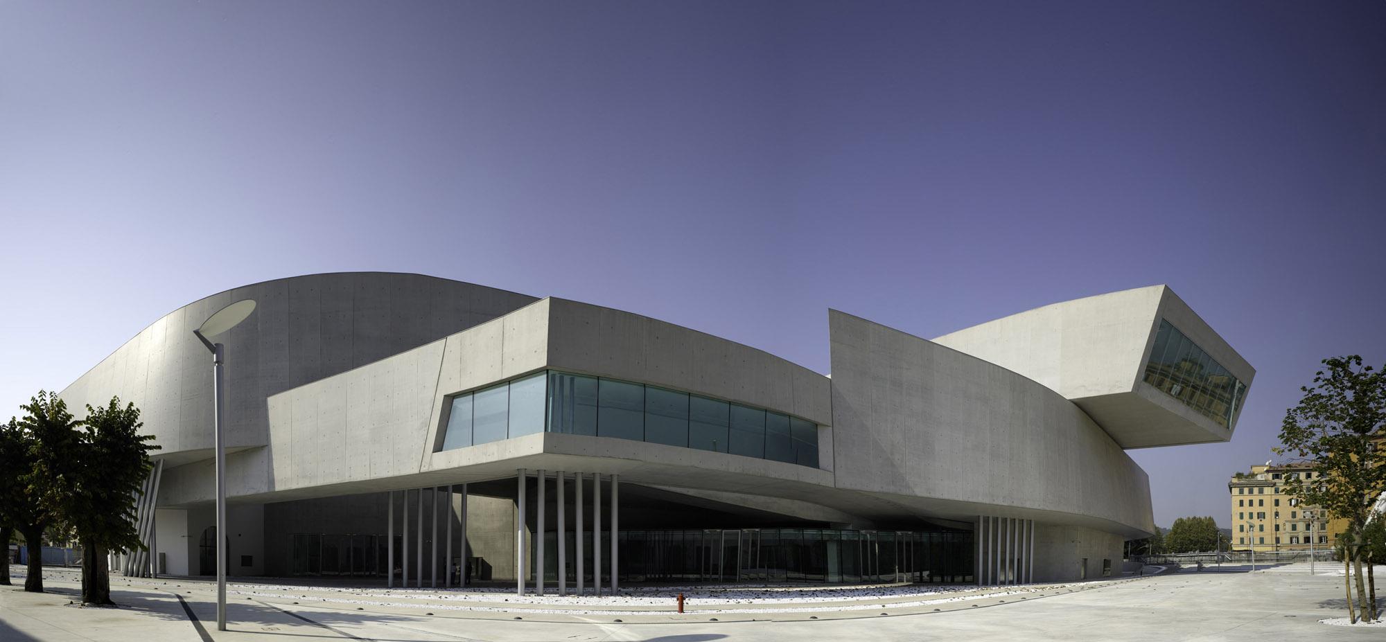 Architecture. Building exterior with metal, glass, and cement, forming both sharp angles and curved surfaces.
This building has at least three distinct stories, but multiple, additional split levels. This wide angle image of the building reveals one side of the building from the street level. One sharp angle juts outward in the middle of the building, on the second story, creating an overhang above the open space below, supported by twelve narrow struts. A wide, geometric window wraps around this sharp angle. On the right end of this side, the building curves around, presumably into the adjacent wall. On top of this curved wall, another geometric, slightly curved structure juts out and creates another overhang, this one on the third story. The overall effect is asymmetrical, yet elegantly balanced.

