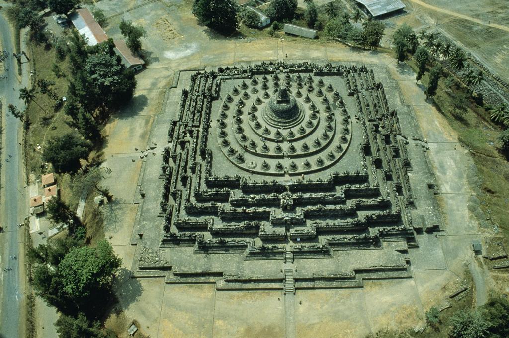 Architecture. Aerial of a building surrounded by trees that is roughly square with subtle curves around the perimeter.
The sides of this square-shaped building appear slightly rounded because of the way each story's perimeter is broken up into different planes. There are at least six levels, each ascending into the next level with less square footage. The structure culminates in a roof with three concentric circles surrounding a dome shape. The aerial shot gives a sense of massive scale.

