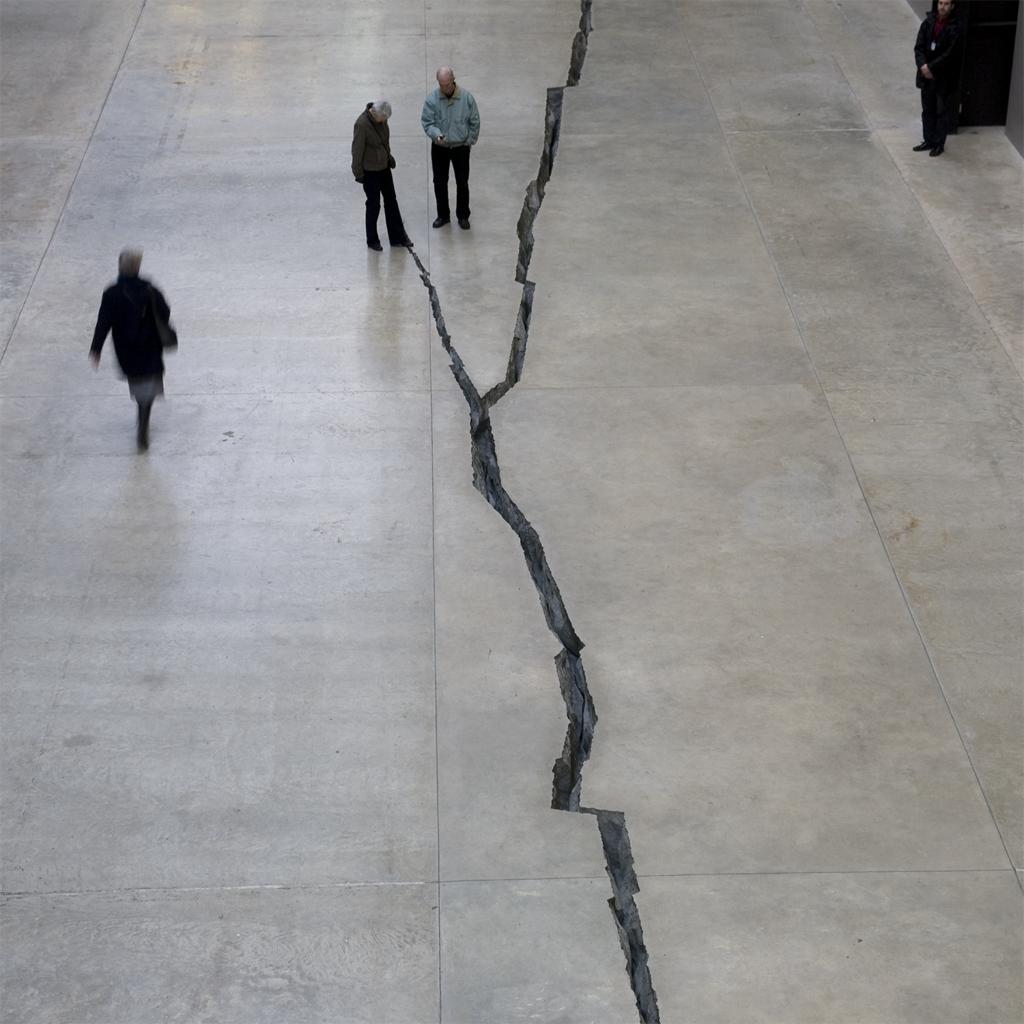 Installation. View of a concrete floor from high above. A crack divides the surface, several people observe the space.
There are four individuals visible in this image, one stands between two branches of the large crack, one stands on the crack itself. Another appears to be walking by, as the other figure stands still, off to the side. The human figures give a sense that the scale of this piece is massive.

