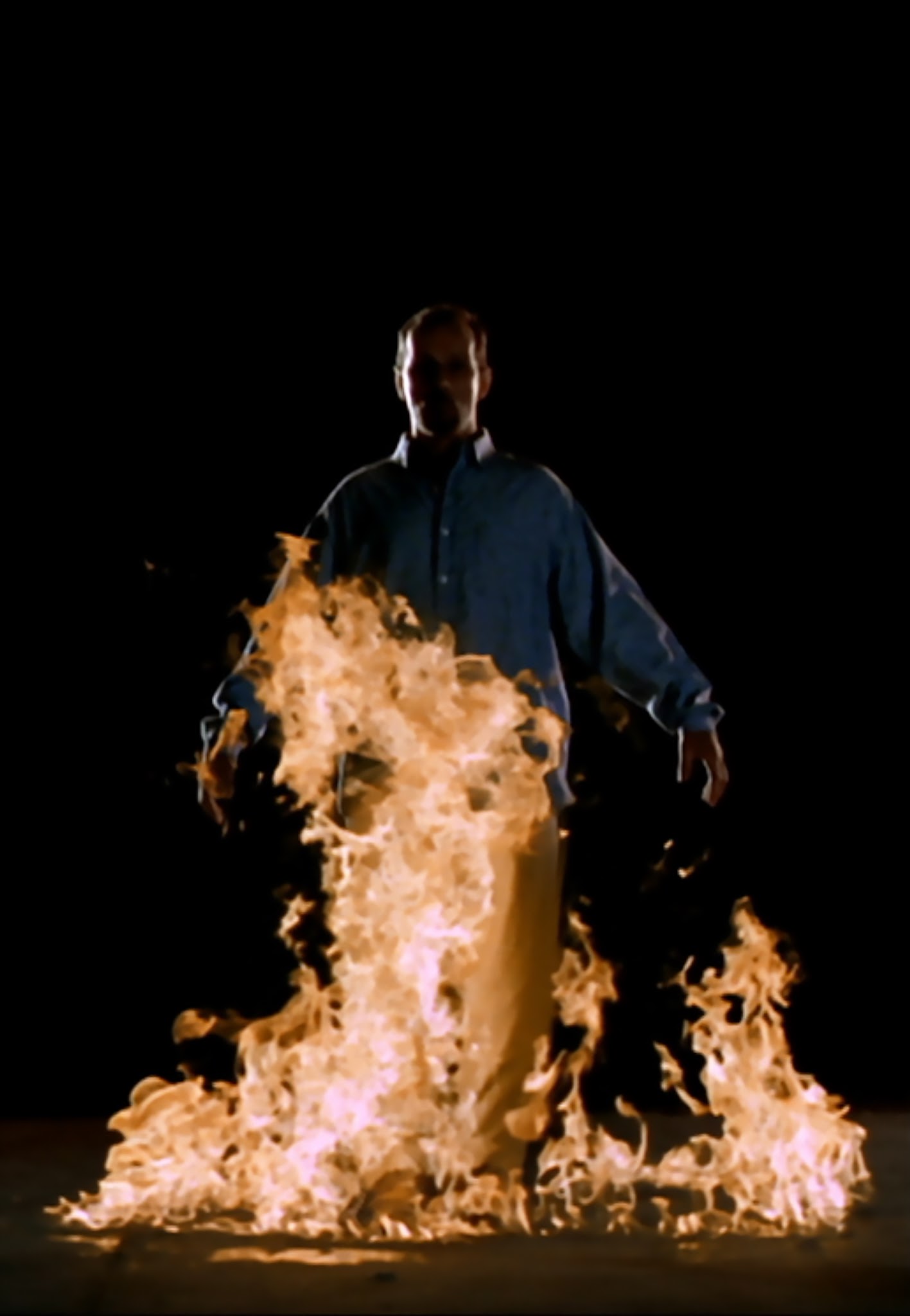Video Still. A man appears to be walking toward the viewer, arms outstretched, his body engulfed in flames.
The man wears a blue shirt, has short hair, and appears very calm, though flames surround him. He appears to be on fire, but not bothered by it.

