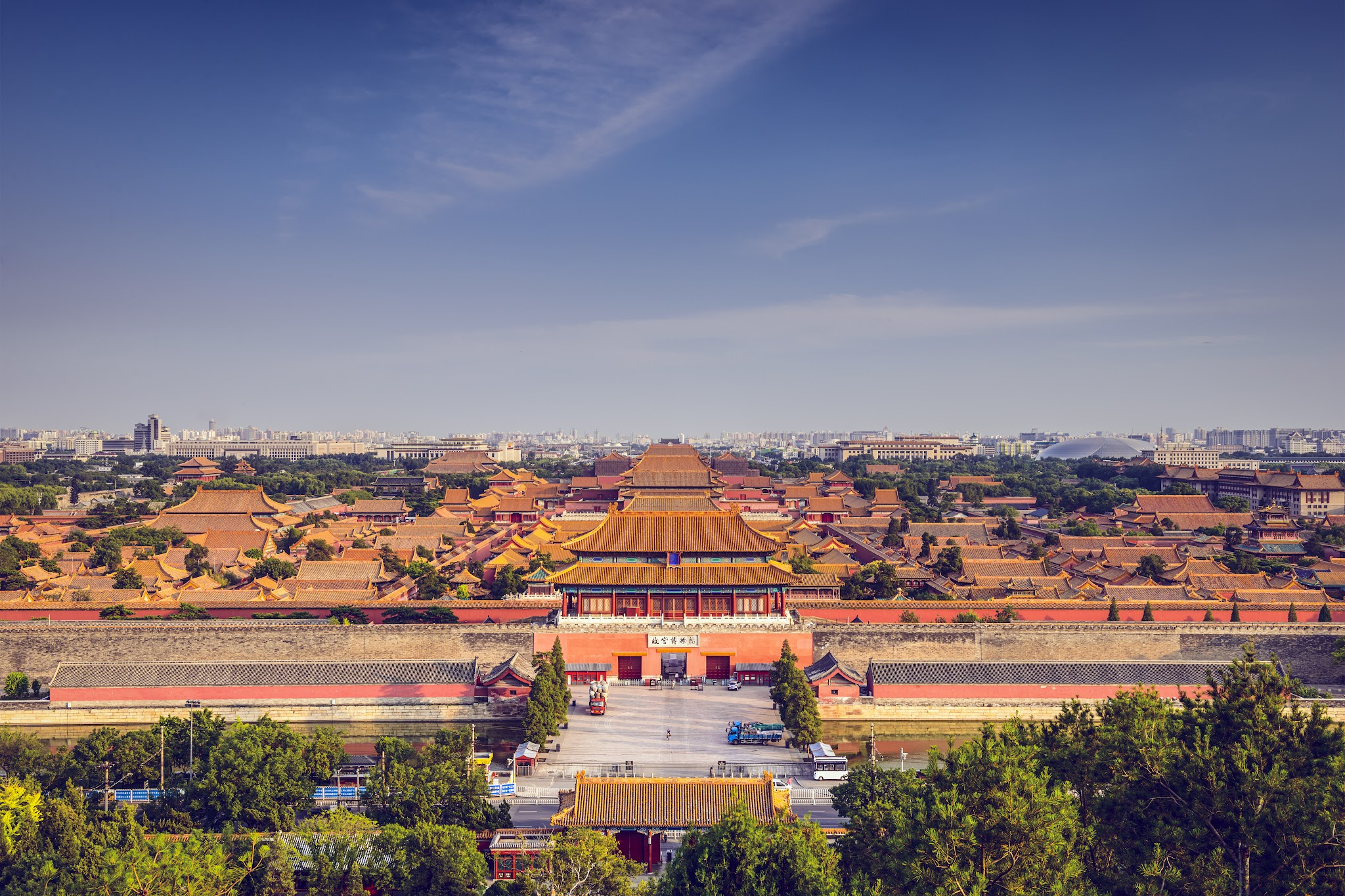 Architecture. Dozens of buildings within a walled complex have dark yellow, flaring roofs.  
From a distance, we look slightly down onto the complex, which extends off both sides of this photograph. A central building appears to be at least three times as tall as any of the others we see here. It has two levels of flat-topped roofs that shallowly curl down and out to each side. These rest on a walkway supported by columns and then on a rectangular base that is several stories high on its own. Trees are interspersed among the other buildings of the complex. A vast city scape is visible in the deep distance beyond.  