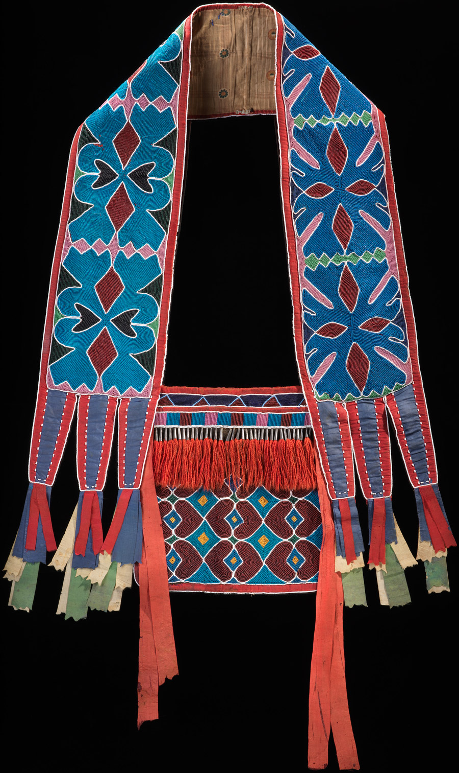 Beadwork on leather. A decorated pouch with a wide strap, adorned with colorful, geometric forms
This piece is decorated in abstract, geometric hues of various reds and blues, with black, white, and gold accents. The overall design has been painted and stitched together, and beads and red tassels have been added.

