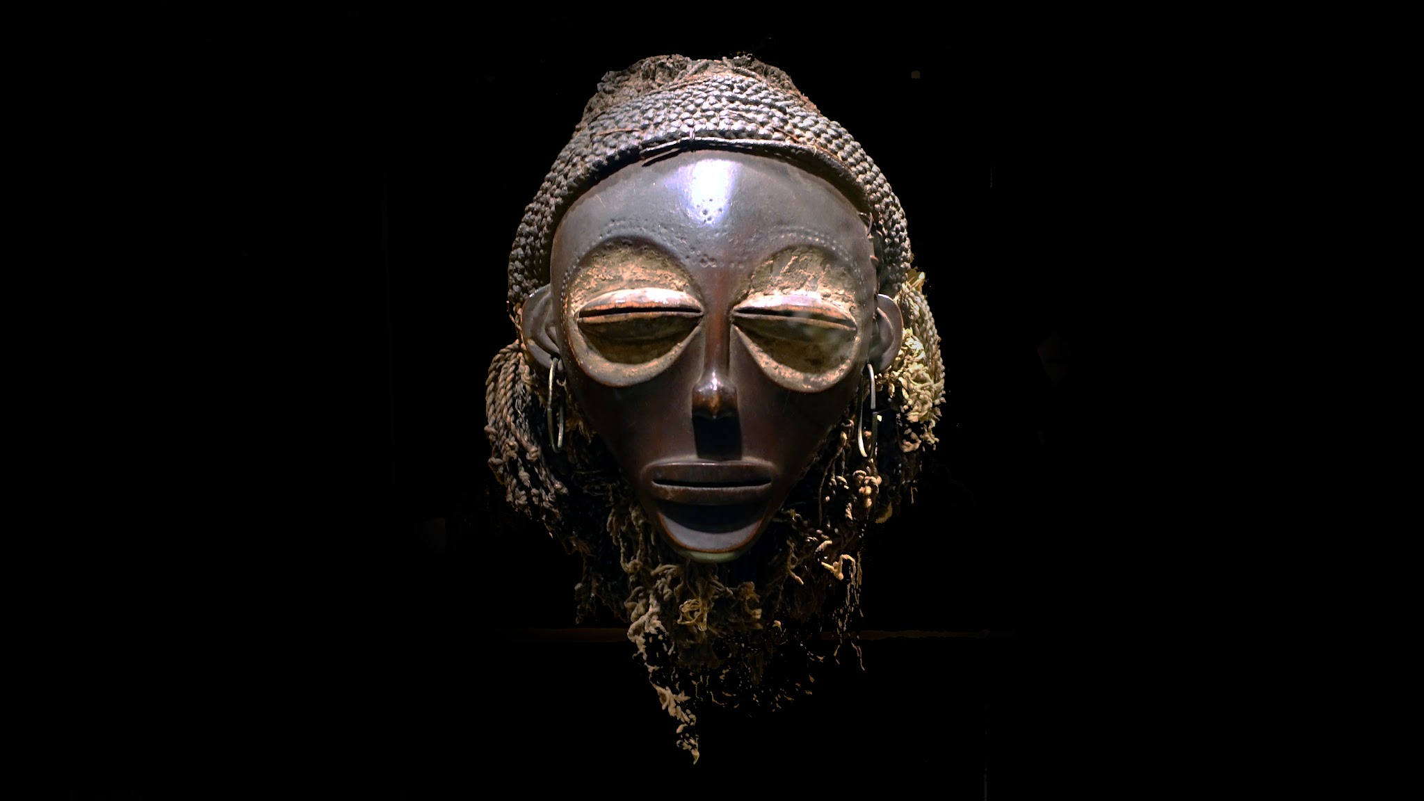 Mask. Smooth, carved wood and fibers, with exaggerated, large eyes and feminine features.
The face of the mask has enlarged, round eye sockets with prominent, mostly-closed, almond-shaped lids. The nose is slender and the balanced face narrows downward to a delicate chin. The braided fiber hair is pulled back away from the face, looped over the crown of the head, and the loose ends dangle to what would be approximately shoulder-length. There are large, hoop earrings in the lobes of the ears.
