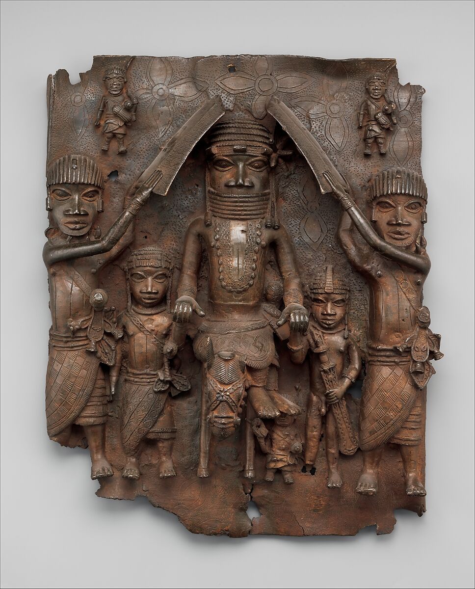 Sculpture. Relief sculpture depicting a large, central figure flanked by two small figures and two medium-sized figures.
This wall plaque contains a total of eight human figures. The largest figure is in the center, with two figures of approximately half its height, immediately to its left and right. There are two slightly larger figures, though not as tall as the central figure, on the far left and right sides of the plaque, and two very small figures that appear to be hovering in the space above them. The final figure is also very small, standing on the ground, partially obscured by the seated, central figure's feet. All of these human figures have similar clothing, features, and exaggerated proportions. They all face directly forwards and wear helmets of varying designs. All are barefoot, with heads of exaggerated size, compared to the rest of their bodies, including shortened legs. They all have very large, wide eyes, staring straight ahead. Several figures hold sword-like weapons, but the two outermost figures appear to hold fan-like objects, as if waving them toward the largest figure. The central figure is the only one wearing a necklace, and a face covering that conceals its mouth and chin.
