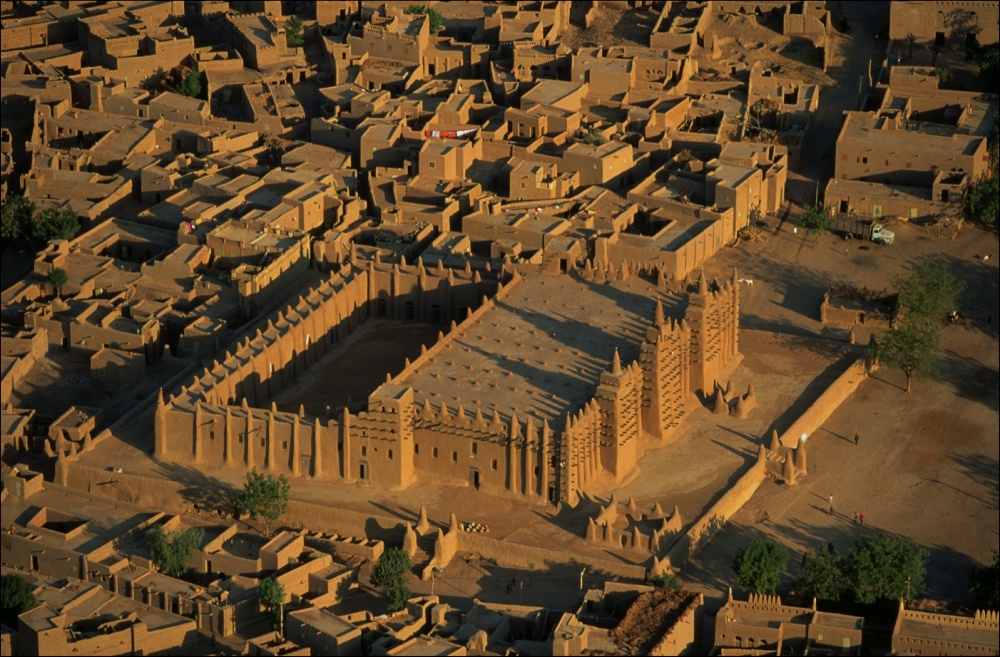 Architecture. A building with one large, open, walled space, and one enclosed space with four minarets across the front.
An aerial view of this building reveals an earthy, monochromatic, sand-colored view of a structure that is much larger than the buildings that surround it. One half of the structure appears to be a walled courtyard, the other half an enclosed space with a flat roof. The minarets are equally spaced across the front of the building. The only visible entryways into the building are small, unobtrusive doors and windows, none of which are located on the facade. Hundreds of short, wooden beams protrude from the side walls in a grid-like pattern.
