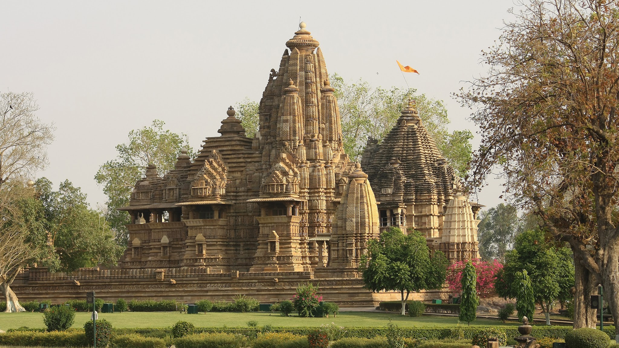 Architecture. This brown sandstone temple is made up of mound-like towers clustered around larger, taller towers.
The entire structure appears to be made of horizontal bands that stretch across all the parts, like the striations of rocks. A central tower rises at the center. Two wider, slightly shorter towers are to each side, and several others nestle around this trio. We look at the temple across a green lawn with manicured bushes.

