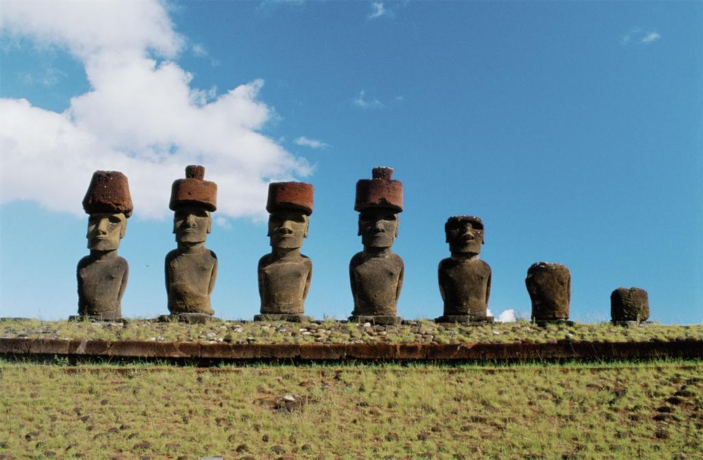 Sculpture. A row of seven massive, stone sculptures, solemn faces and wide eyes. The five on the left kneel, hands behind their backs. The two on right are missing ther top portions.Five of the stone sculptures of human-like figures appear to be kneeling, hands behind their backs. All of their faces wear a calm, solemn expression, and four of the five figures wear a large, circular hat. The figures kneel in an upright, regal manner, staring outwards, with a bright blue sky behind them. To th right, one of the sculptures is missing the head and the other is missing the head and part of the body. 