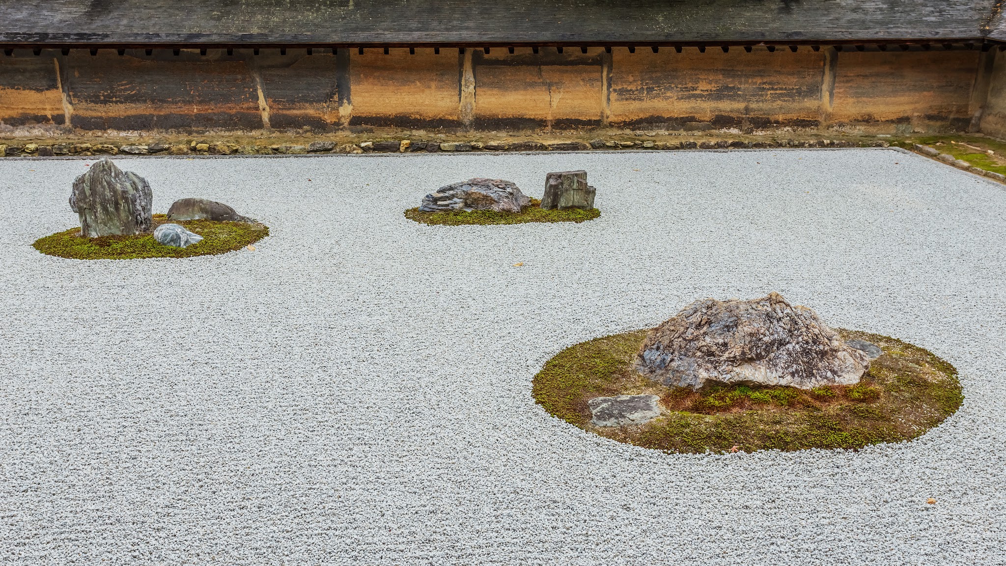Rock garden. Clusters of gray rocks are arranged on three grassy spots amid a field of raked gray gravel.
Lines raked into the gravel encircle the grass and create waves between them. The rocks are different sizes and shapes, with some being low and wide and others tall and narrow, like teeth. The clay wall enclosing this view is charcoal gray in some areas and a warm orange in others.

