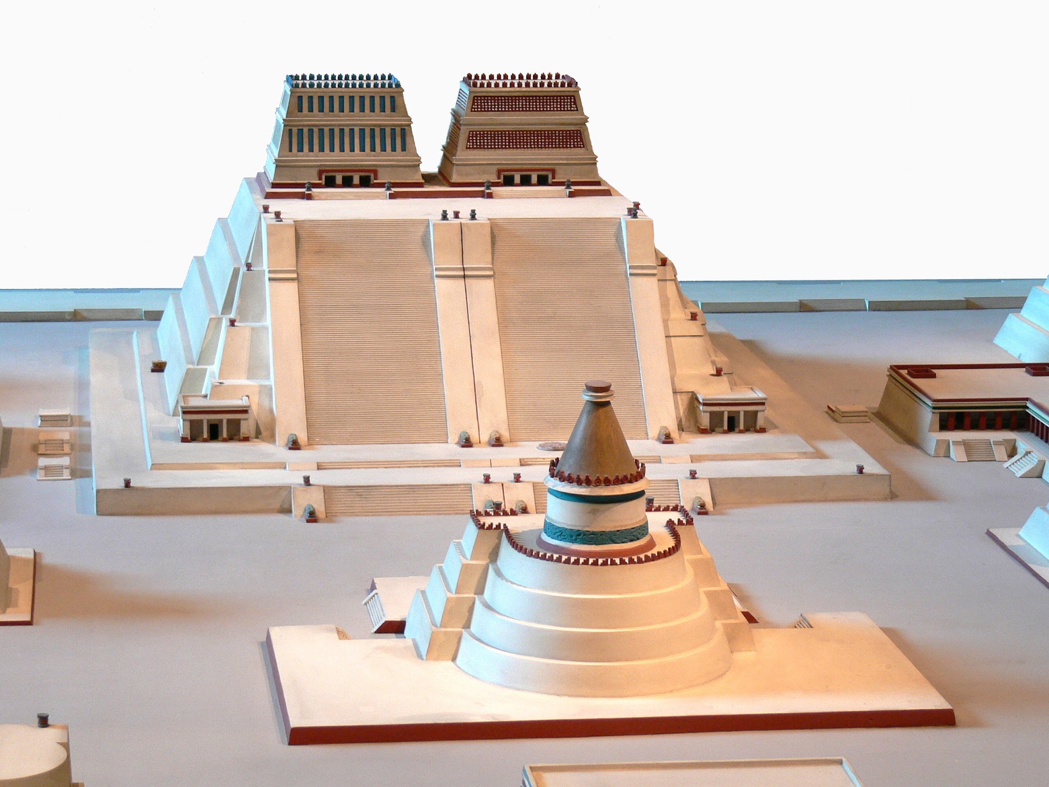 Architecture. Virtual reconstruction of a building complex, one building conical shaped, the other pyramidal shaped. This digital reconstruction shows us a rendering of a building complex that has two main, visible structures. In the background there is a pyramid-shaped building. The top several stories of the pyramidal building are divided into two separate spaces with elaborate roofs, of equal size but slightly different design. Two separate sets of stairs lead to each of these topmost structures. The building in the foreground is conical, with each level rising upwards in diminishing, concentric rings. It is capped by a painted, decorated, pointed roof that completes the conical pattern. 
